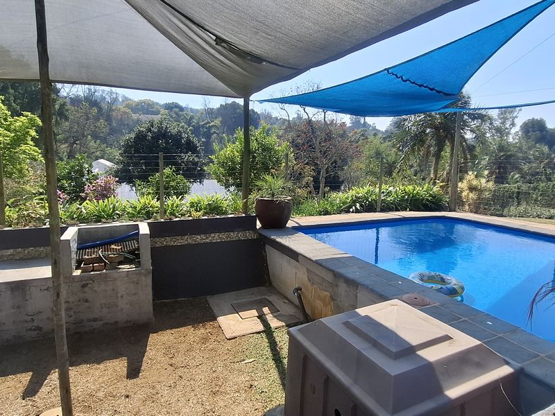 Outdoor pool area featuring a built-in braai, shade sail coverage, and a swimming pool set against a landscaped garden backdrop.