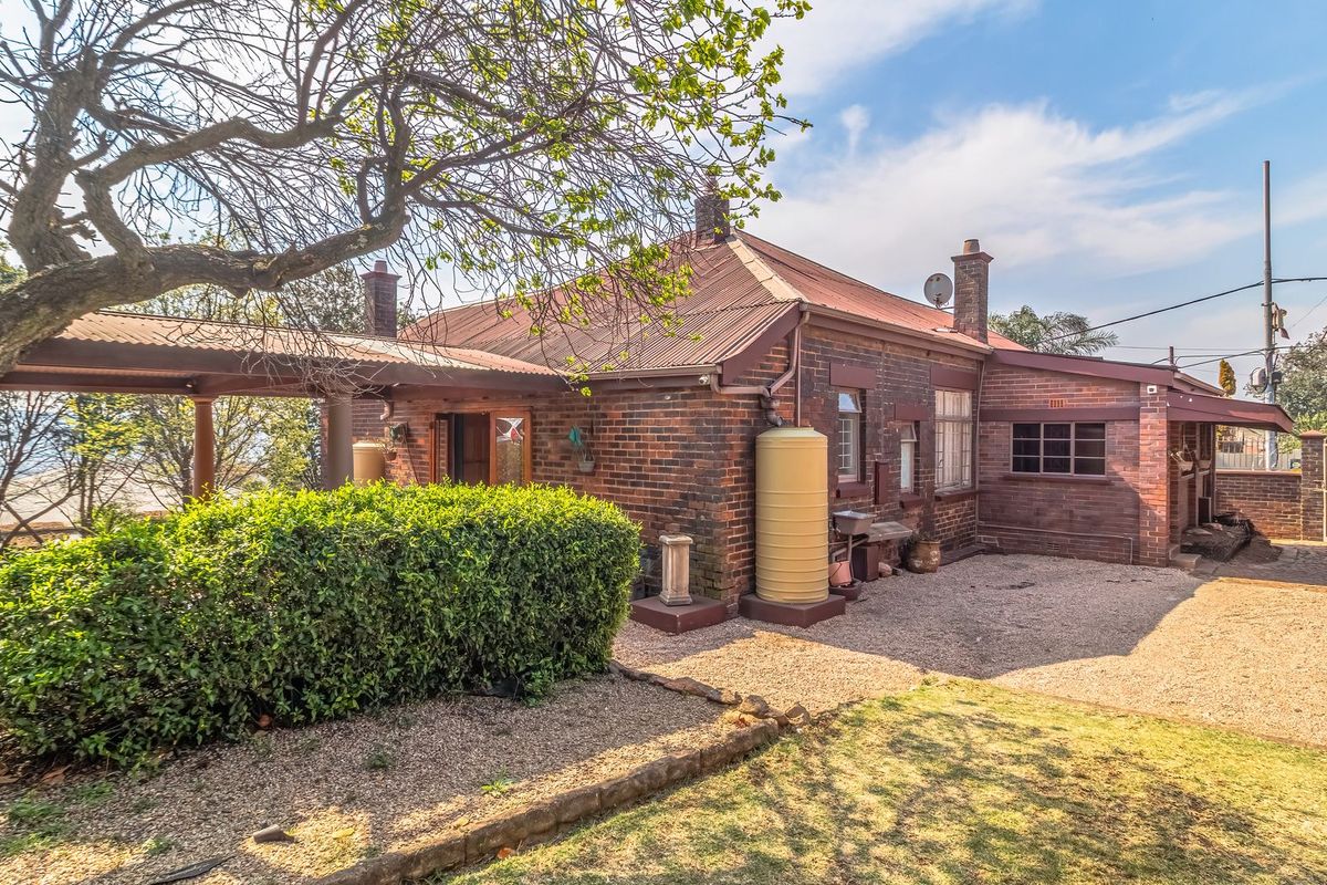 Rear view of a traditional brick house featuring a corrugated metal roof, covered patio area, and a gravel driveway with established garden surroundings.