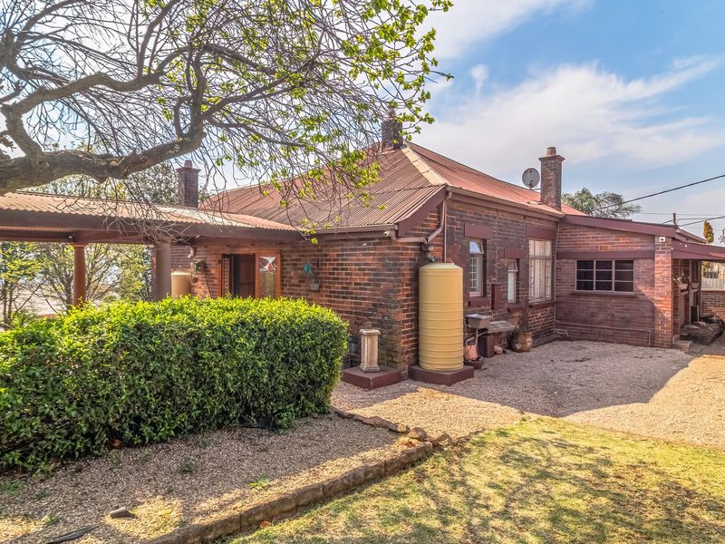 Rear view of a traditional brick house featuring a corrugated metal roof, covered patio area, and a gravel driveway with established garden surroundings.