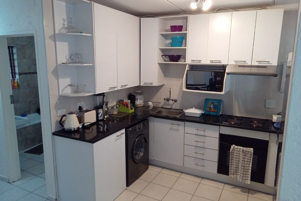 Compact kitchen featuring white cabinetry, black countertops, built-in oven, electric hob, extractor fan, and tiled flooring with an adjacent bathroom view.