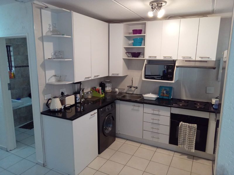Compact kitchen featuring white cabinetry, black countertops, built-in oven, electric hob, extractor fan, and tiled flooring with an adjacent bathroom view.