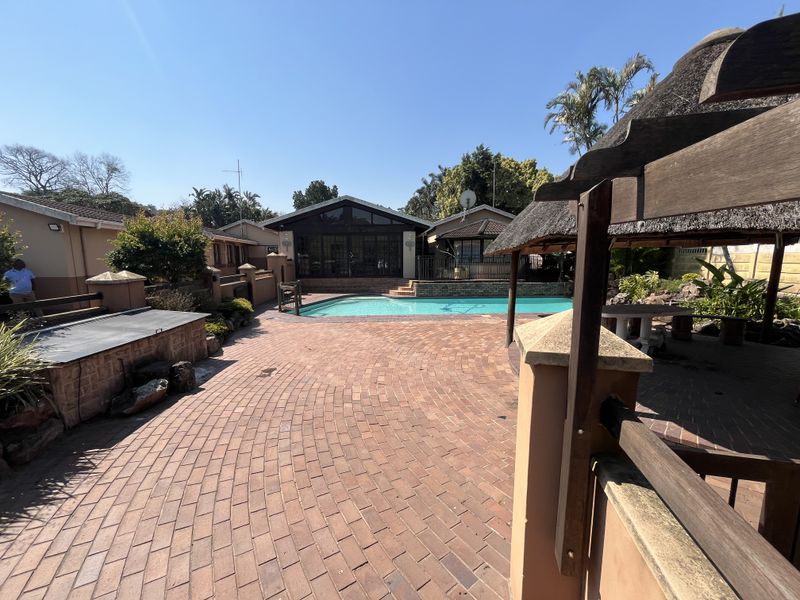 Outdoor pool area featuring brick paving, a swimming pool, and a thatched gazebo structure in a landscaped garden setting.