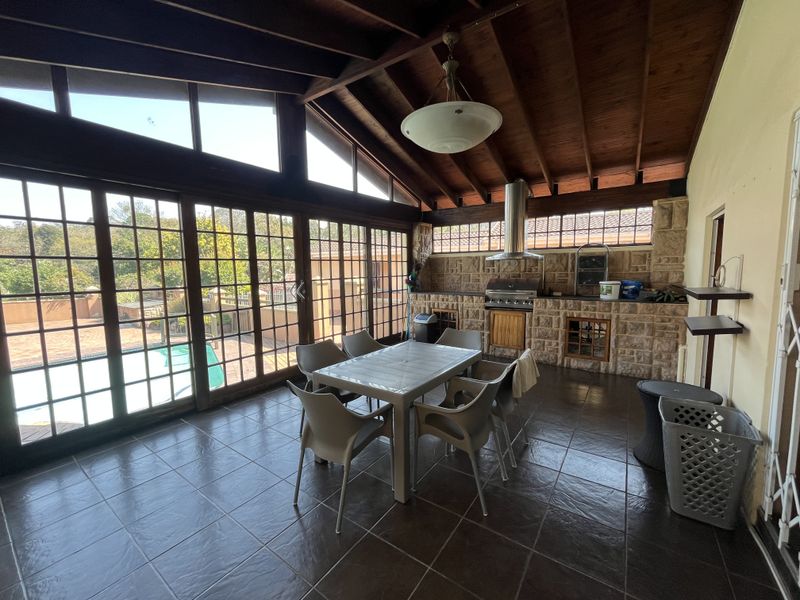 Spacious covered patio featuring a stone-clad built-in braai, exposed timber ceiling, and large glass sliding doors overlooking the pool area.
