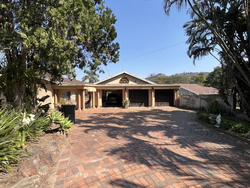 Front elevation of the property featuring a circular brick paved driveway, double garage, covered carport area, and established garden surroundings.