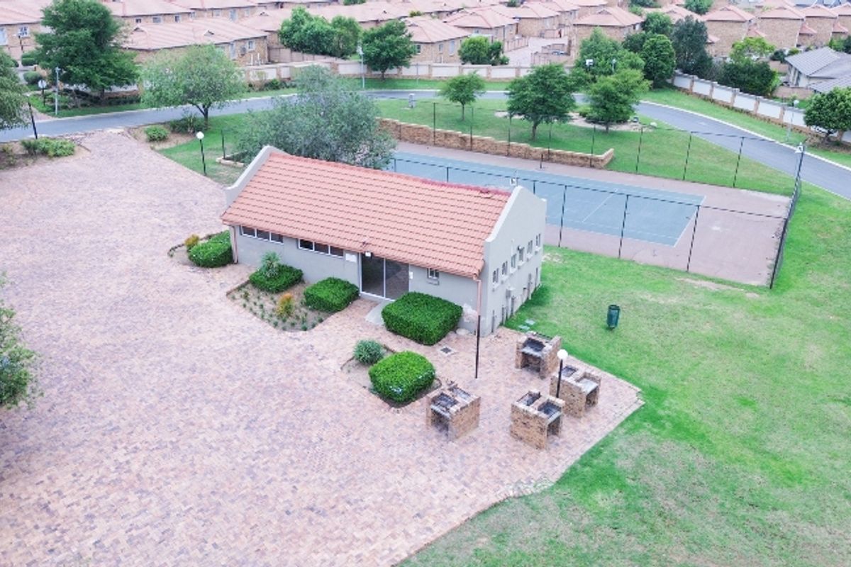 Aerial view of the complex clubhouse featuring a tennis court, paved outdoor entertainment area with built-in braai facilities, and well-maintained communal gardens.