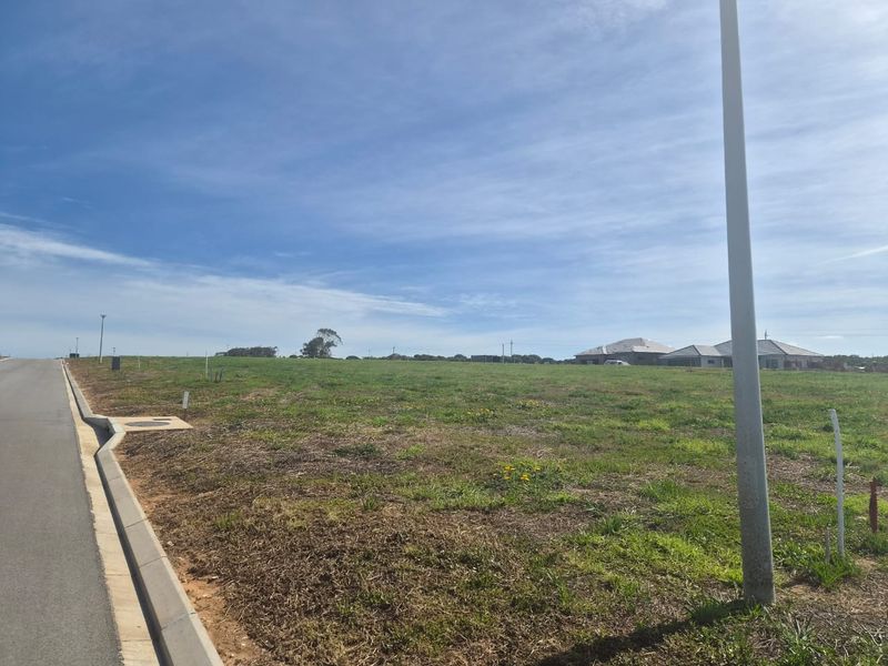 A vacant residential land parcel featuring a level grassed surface, adjacent to a paved road with established curb and utility infrastructure.
