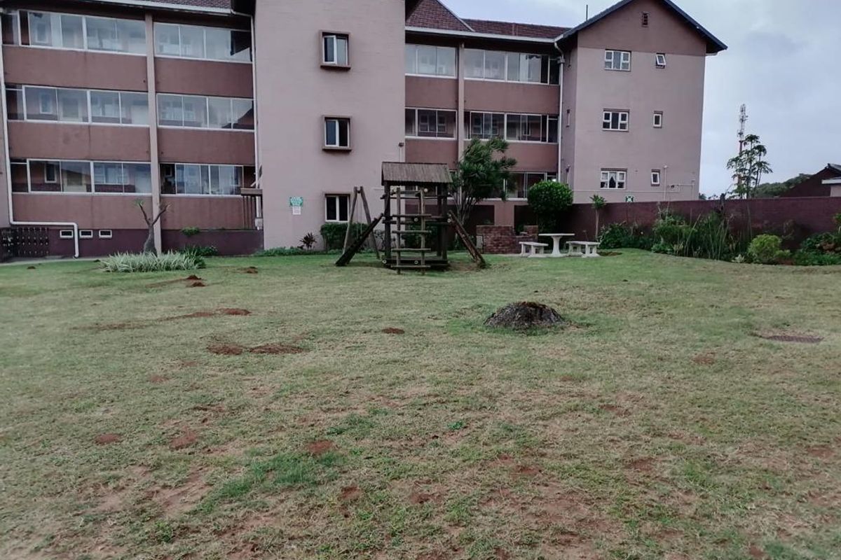 A view of the communal rear garden area featuring a lawn, playground equipment, and a brick boundary wall at the back of the apartment complex.