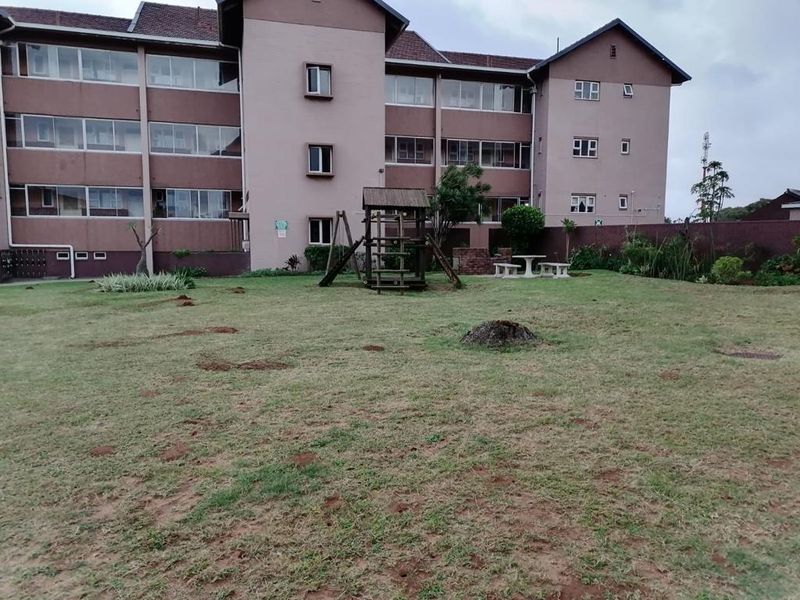 A view of the communal rear garden area featuring a lawn, playground equipment, and a brick boundary wall at the back of the apartment complex.