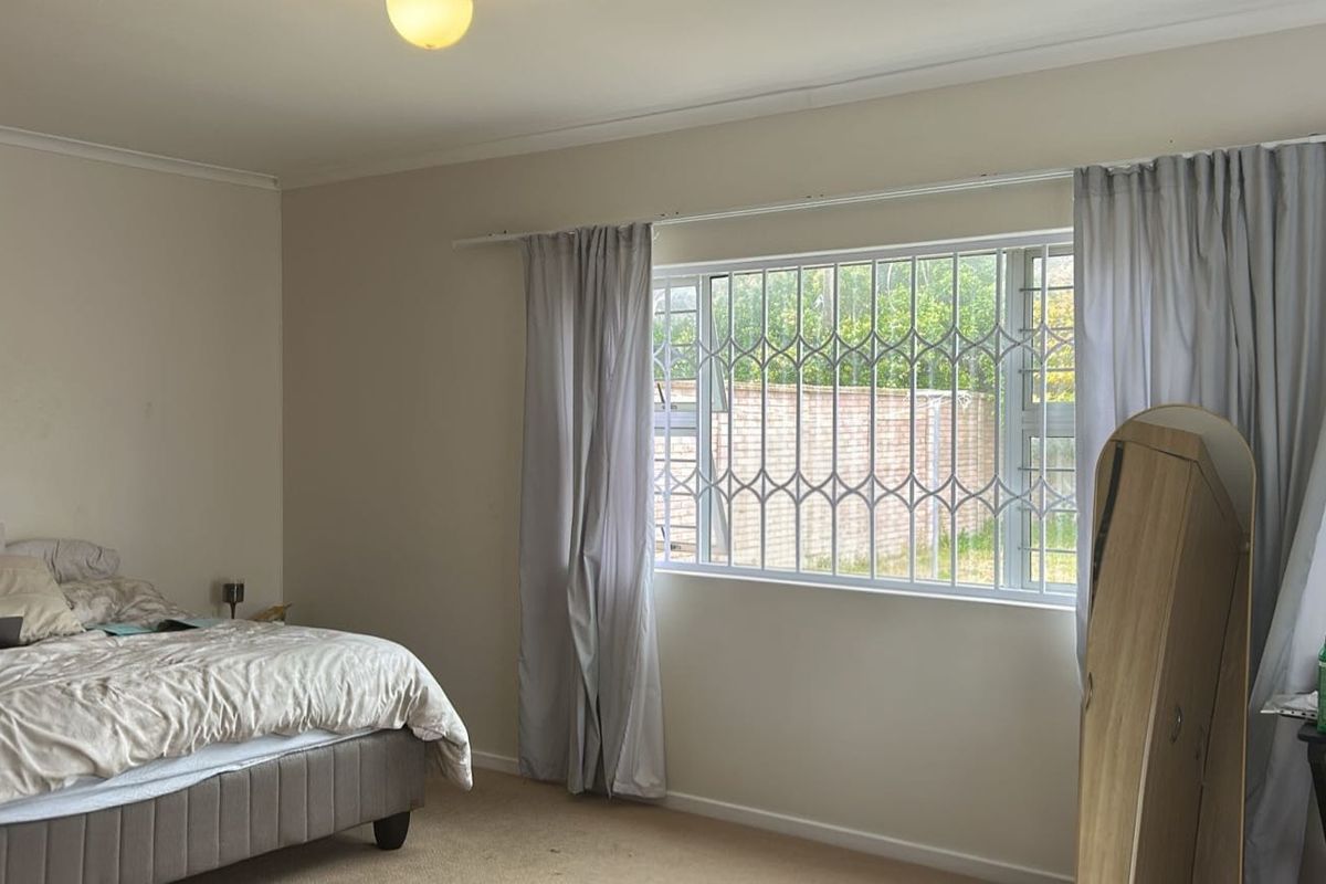 A spacious bedroom featuring neutral walls, carpeted flooring, and a window fitted with a security gate for added safety.