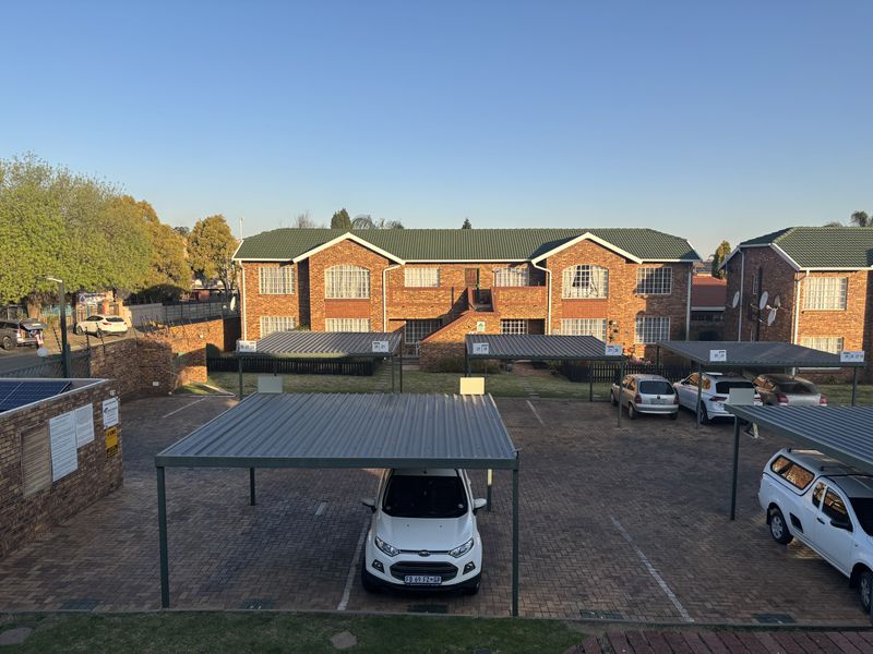An elevated view of an apartment complex featuring a brick facade, tiled roof, and multiple covered parking bays on a paved driveway.