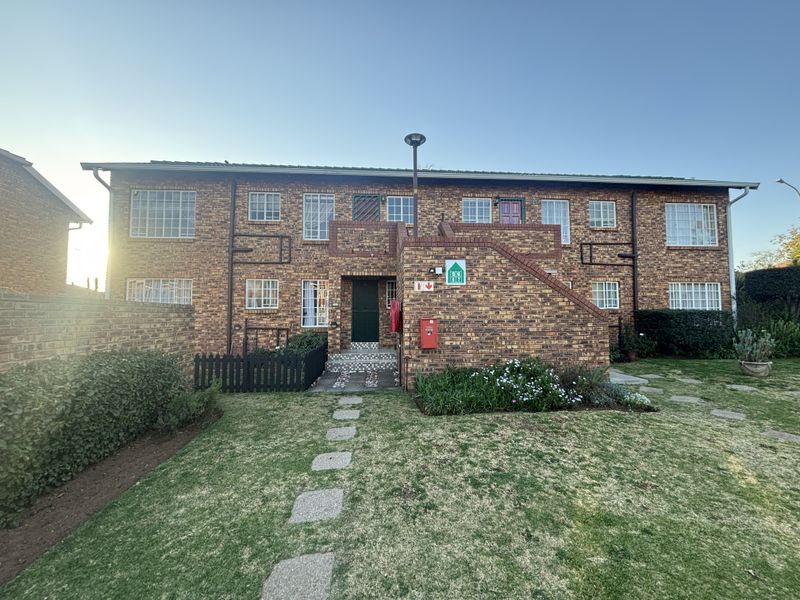 Front view of a two-story face brick apartment building featuring an external staircase, security bars on windows, and a well-maintained garden with a paved walkway.