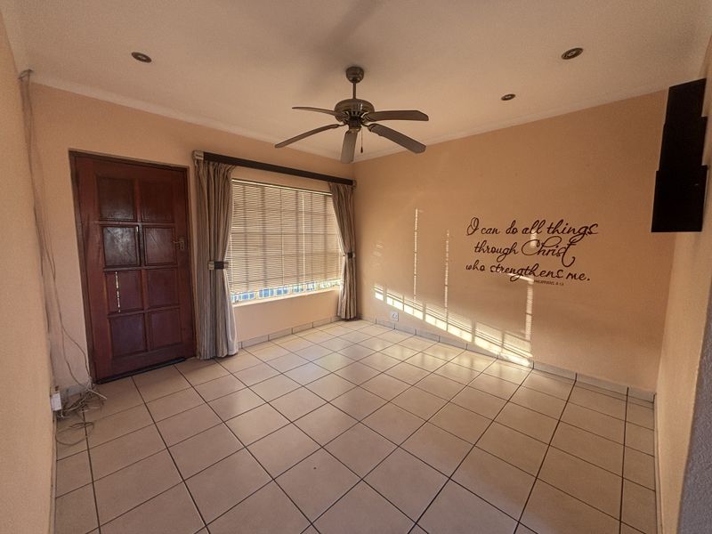 A bright, empty living room featuring tiled flooring, a ceiling fan, recessed lighting, and a timber entrance door with venetian blinds.