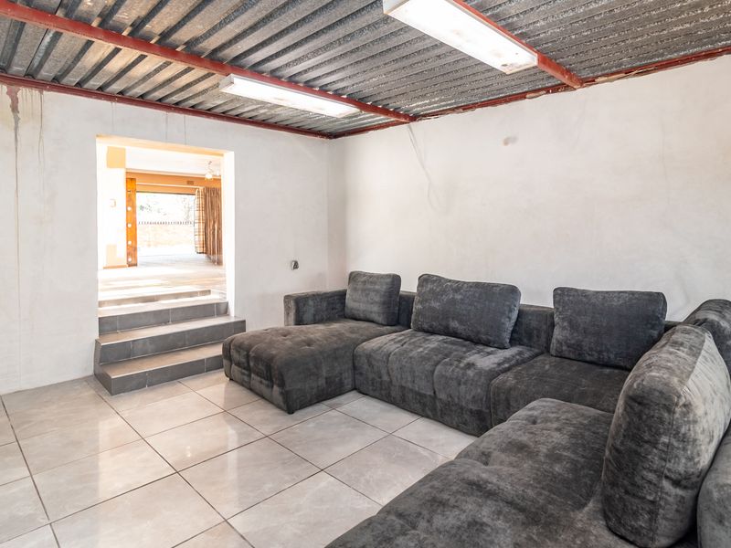 A living area featuring tiled floors and an exposed corrugated metal ceiling, leading to an adjacent space via a small set of stairs.