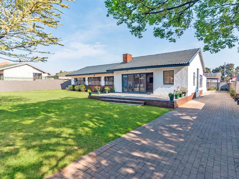 Rear view of the house featuring a well-maintained lawn, paved driveway, raised patio area, and sliding glass doors leading into the home.