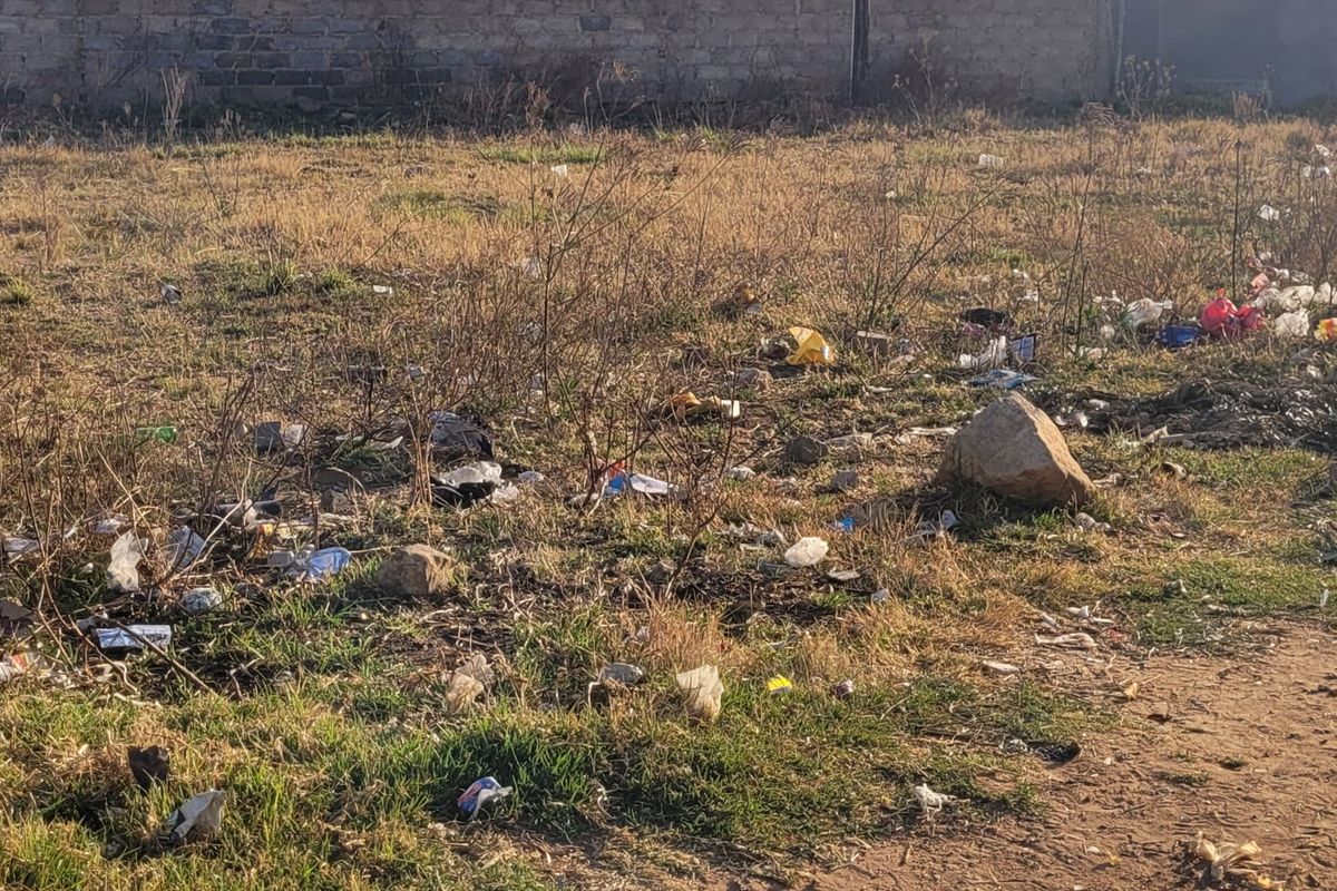 A vacant residential plot featuring dry grass, scattered debris, and a perimeter boundary wall.