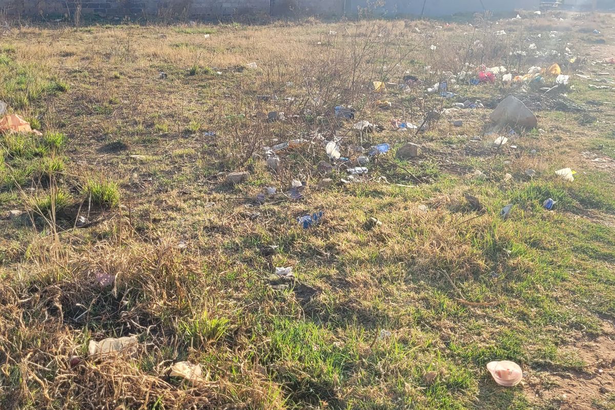A vacant residential plot featuring unmaintained grass and significant accumulation of litter and debris across the site.