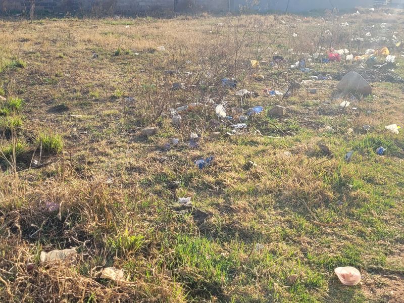 A vacant residential plot featuring unmaintained grass and significant accumulation of litter and debris across the site.