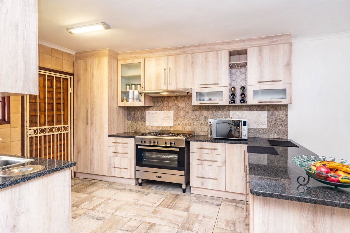 Modern kitchen featuring light wood cabinetry, granite countertops, a gas stove with extractor fan, and tiled flooring with an adjacent security gate.