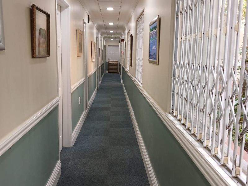 A long, well-lit apartment hallway featuring wainscoting, carpeted flooring, and a secure metal gate along the side wall.