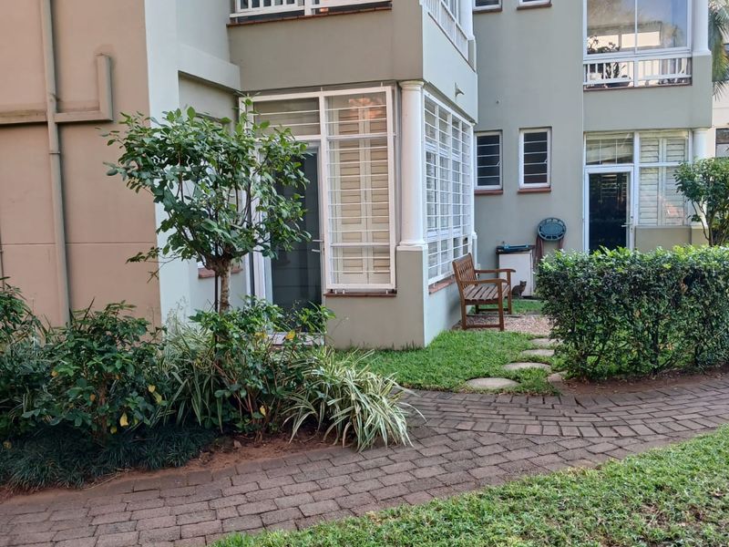A view of the apartment building's rear exterior, featuring a paved walkway, landscaped garden, and enclosed balcony areas with security bars.