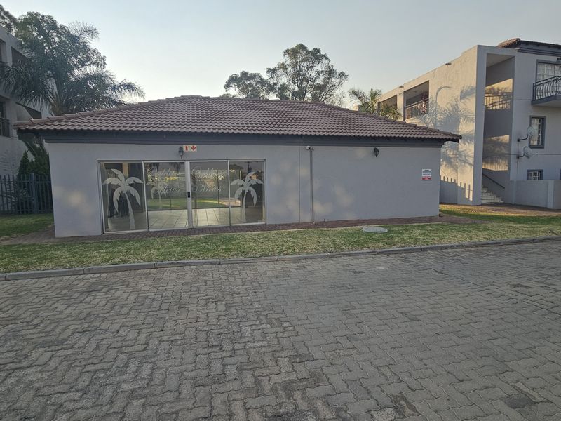 A view of the complex clubhouse building featuring a tiled roof and sliding glass door entrance, situated within a paved townhouse estate.