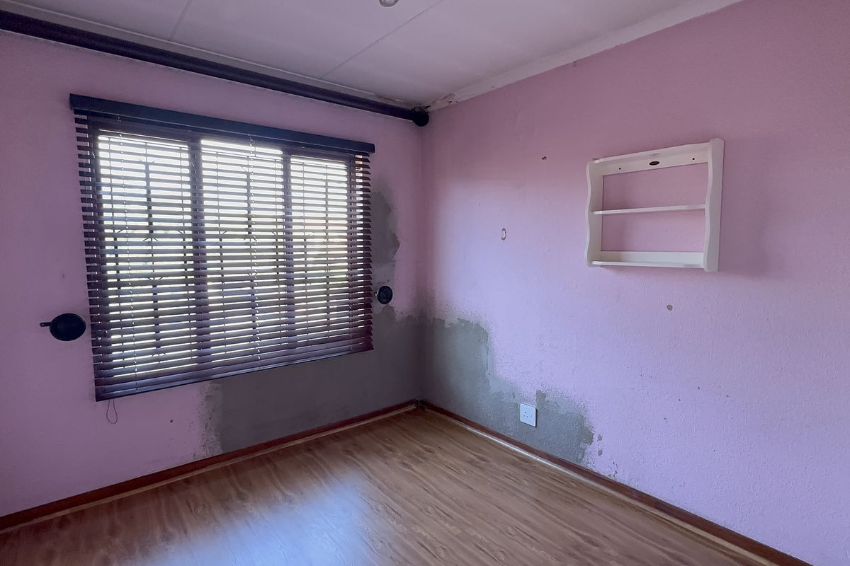An empty bedroom featuring laminate flooring, venetian blinds, and a built-in wall shelf, currently showing visible wall plaster damage and signs of moisture.