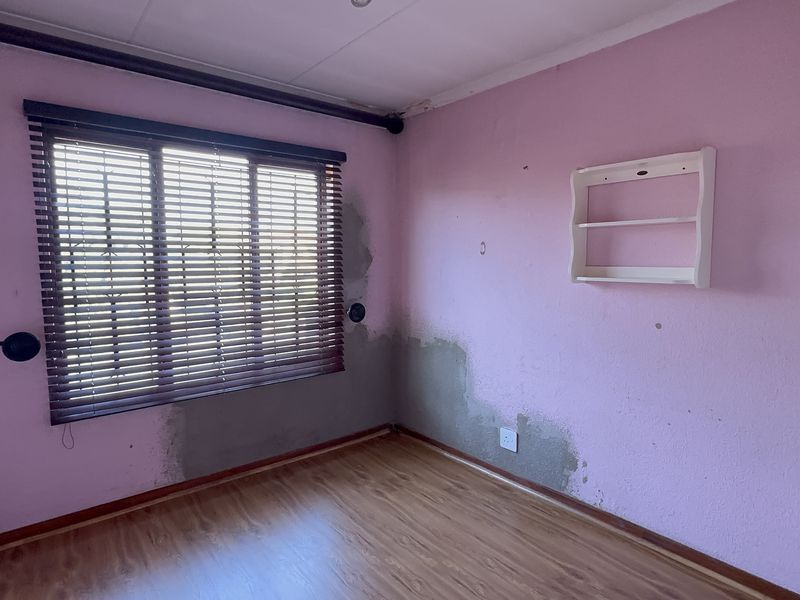An empty bedroom featuring laminate flooring, venetian blinds, and a built-in wall shelf, currently showing visible wall plaster damage and signs of moisture.