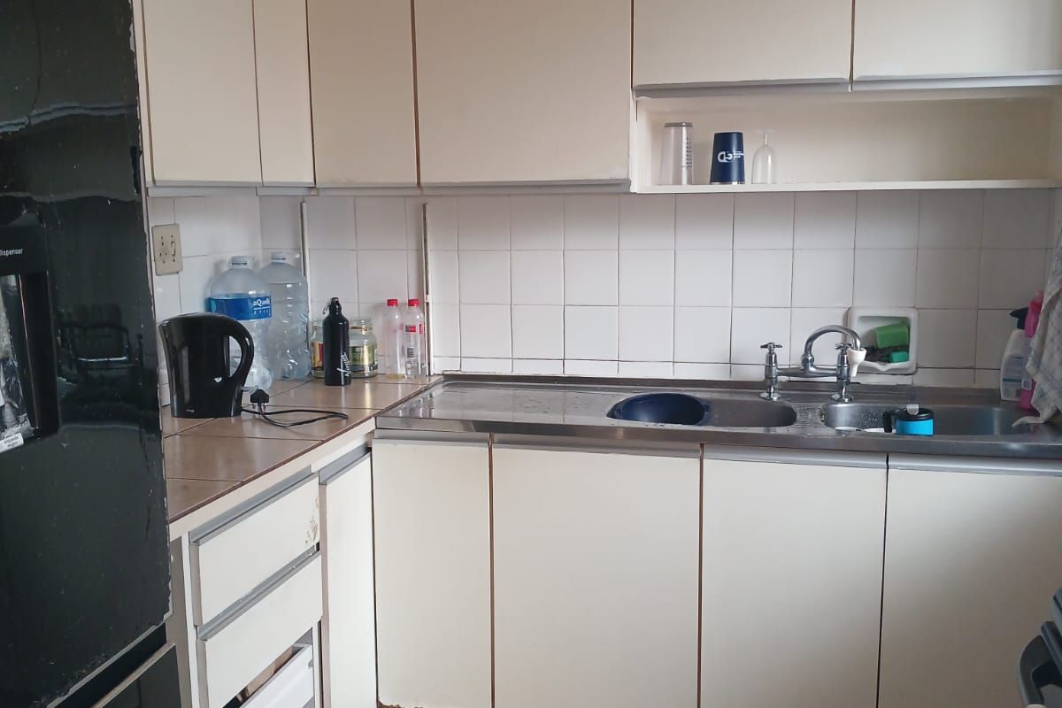 A compact kitchen featuring white laminate cabinetry, a stainless steel sink, tiled backsplash, and ceramic floor tiles, showing signs of wear.