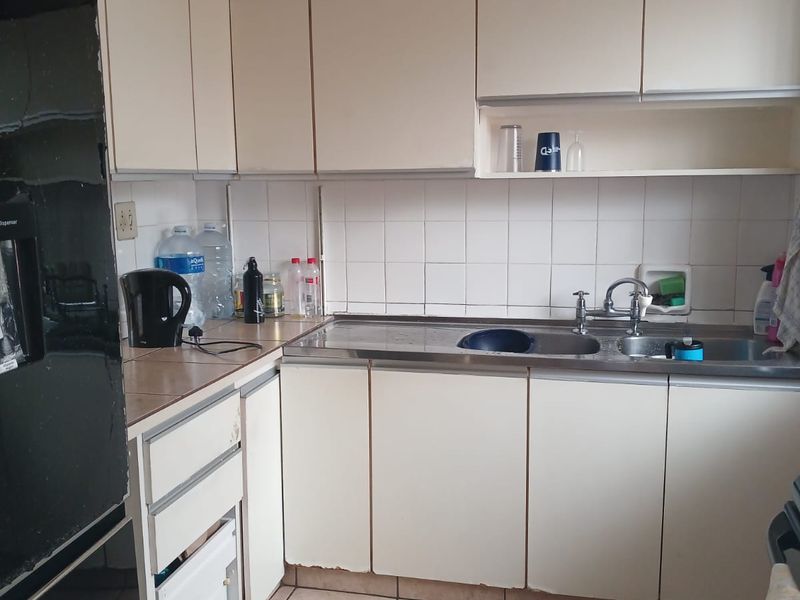 A compact kitchen featuring white laminate cabinetry, a stainless steel sink, tiled backsplash, and ceramic floor tiles, showing signs of wear.