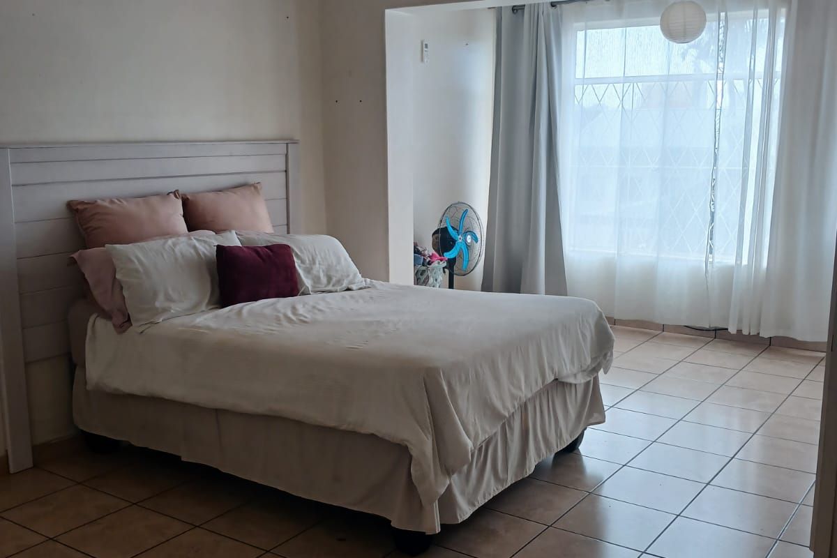 A spacious bedroom featuring tiled flooring, a built-in headboard, and a large window with security bars, offering a functional and well-lit living space.