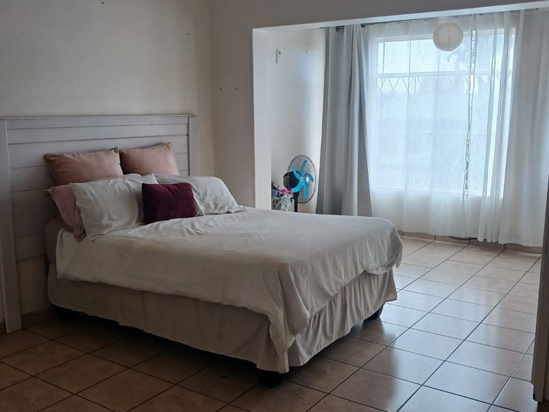 A spacious bedroom featuring tiled flooring, a built-in headboard, and a large window with security bars, offering a functional and well-lit living space.