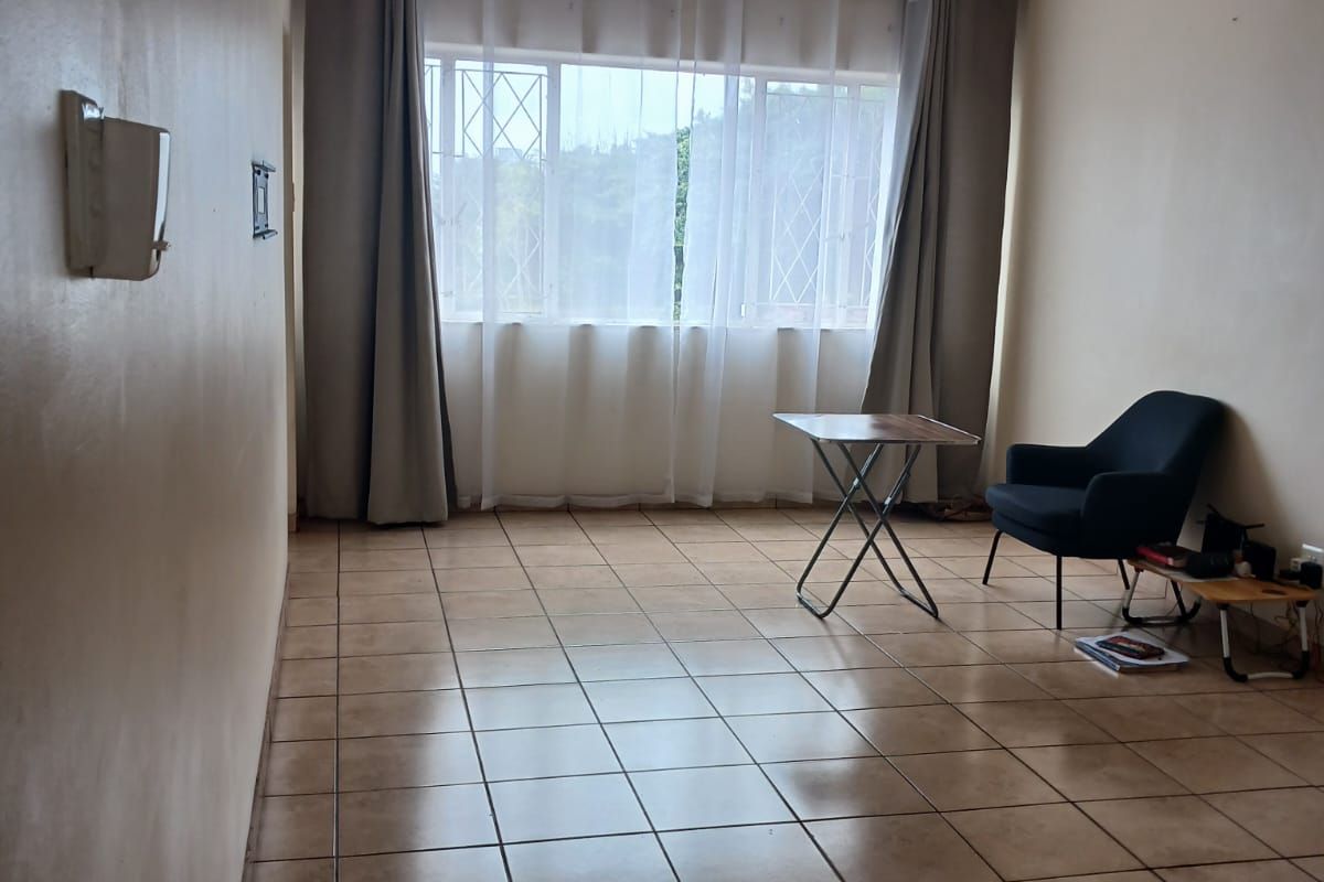 A spacious, neutrally painted living area featuring ceramic floor tiles, a ceiling fan, and large windows with security gates.