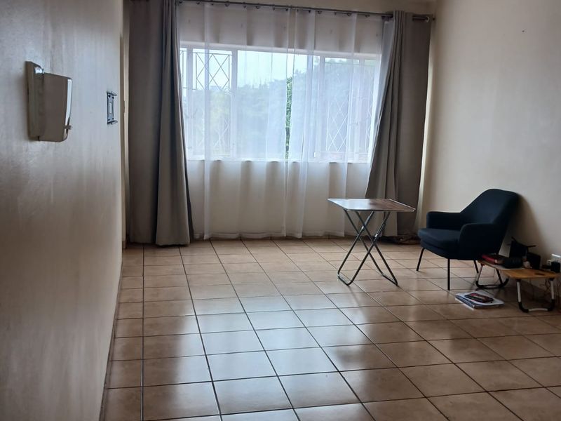 A spacious, neutrally painted living area featuring ceramic floor tiles, a ceiling fan, and large windows with security gates.