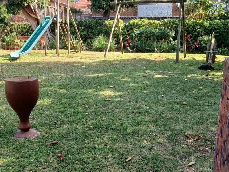 A communal garden area featuring a lawn, mature trees, and a perimeter wall with security fencing, situated within an apartment complex.