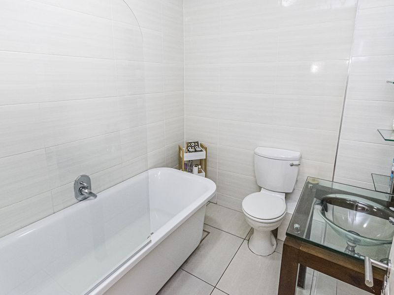 A clean, modern bathroom featuring white wall tiles, a bathtub, a toilet, and a glass vessel sink vanity with integrated shelving.