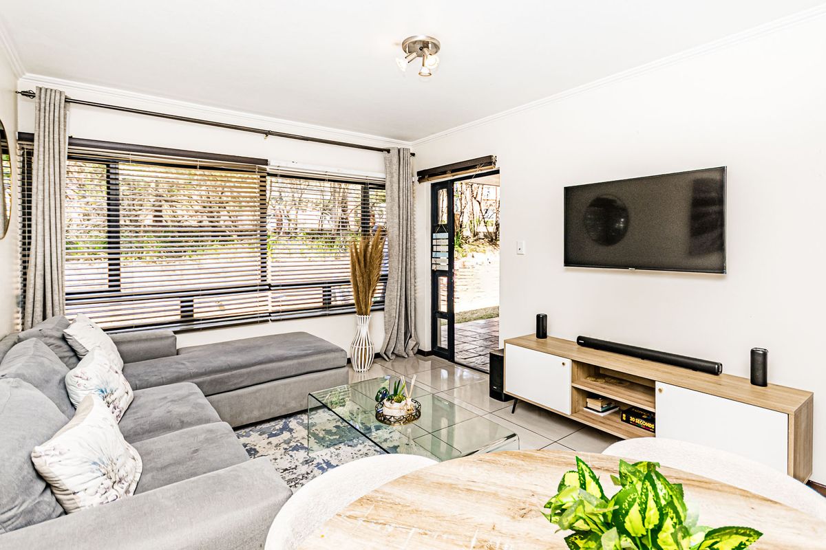 A bright and well-maintained living room featuring tiled flooring, large windows with blinds, and direct access to an outdoor area through a sliding glass door.