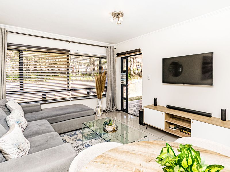 A bright and well-maintained living room featuring tiled flooring, large windows with blinds, and direct access to an outdoor area through a sliding glass door.