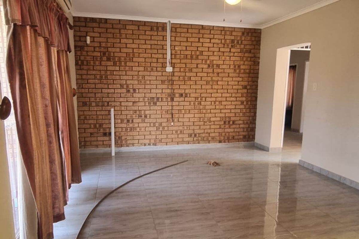 Spacious living area featuring a prominent exposed brick wall, neutral tiled flooring, ceiling fan, and wall-mounted air conditioning unit.
