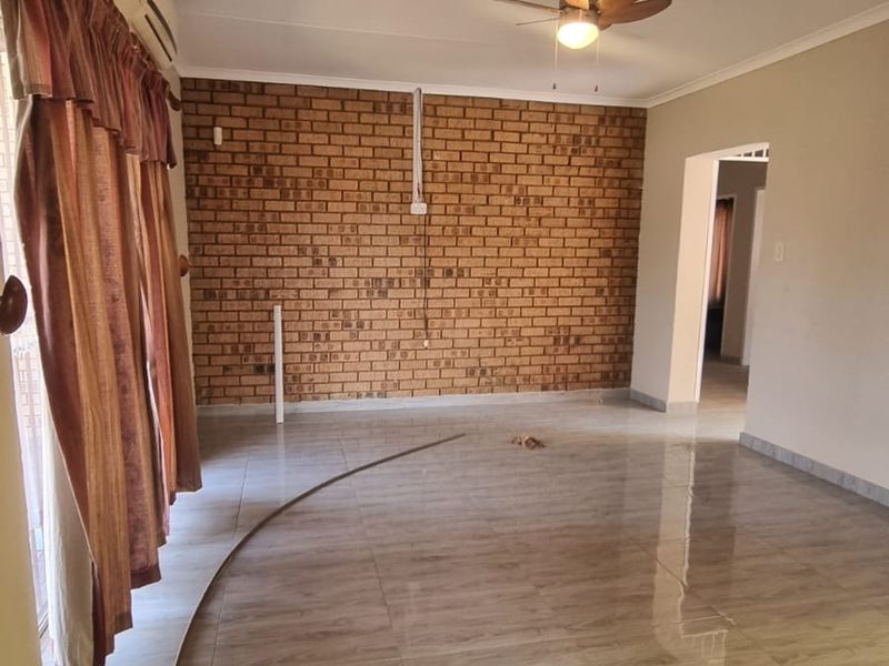 Spacious living area featuring a prominent exposed brick wall, neutral tiled flooring, ceiling fan, and wall-mounted air conditioning unit.