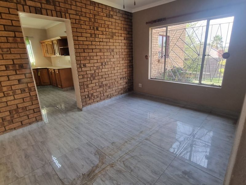 An empty living room featuring an exposed brick wall, tiled flooring, and a large window with security bars, leading into the kitchen area.