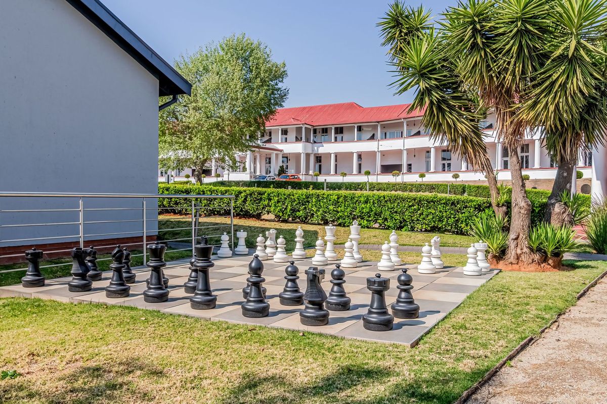 A well-maintained outdoor courtyard area featuring a large garden chess set, paved flooring, and manicured hedges set against a multi-story apartment building.