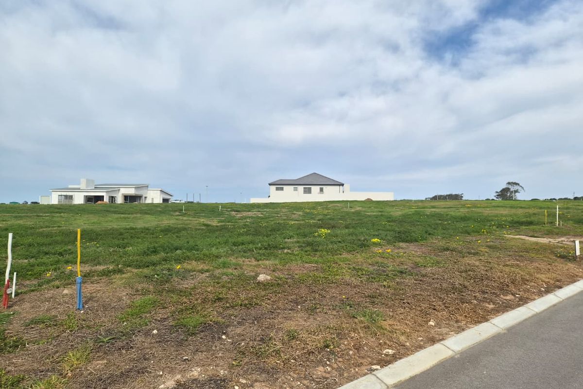 A vacant residential plot of land featuring a grassy landscape with nearby established homes and paved road access.
