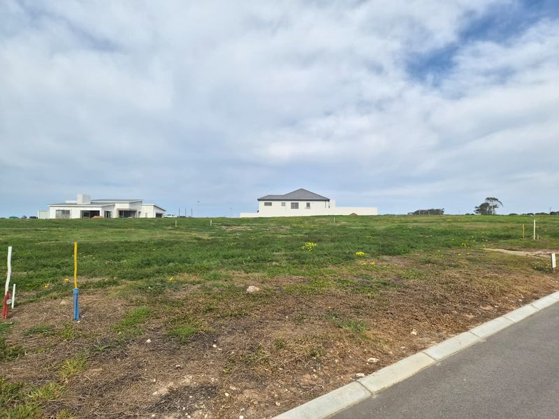 A vacant residential plot of land featuring a grassy landscape with nearby established homes and paved road access.