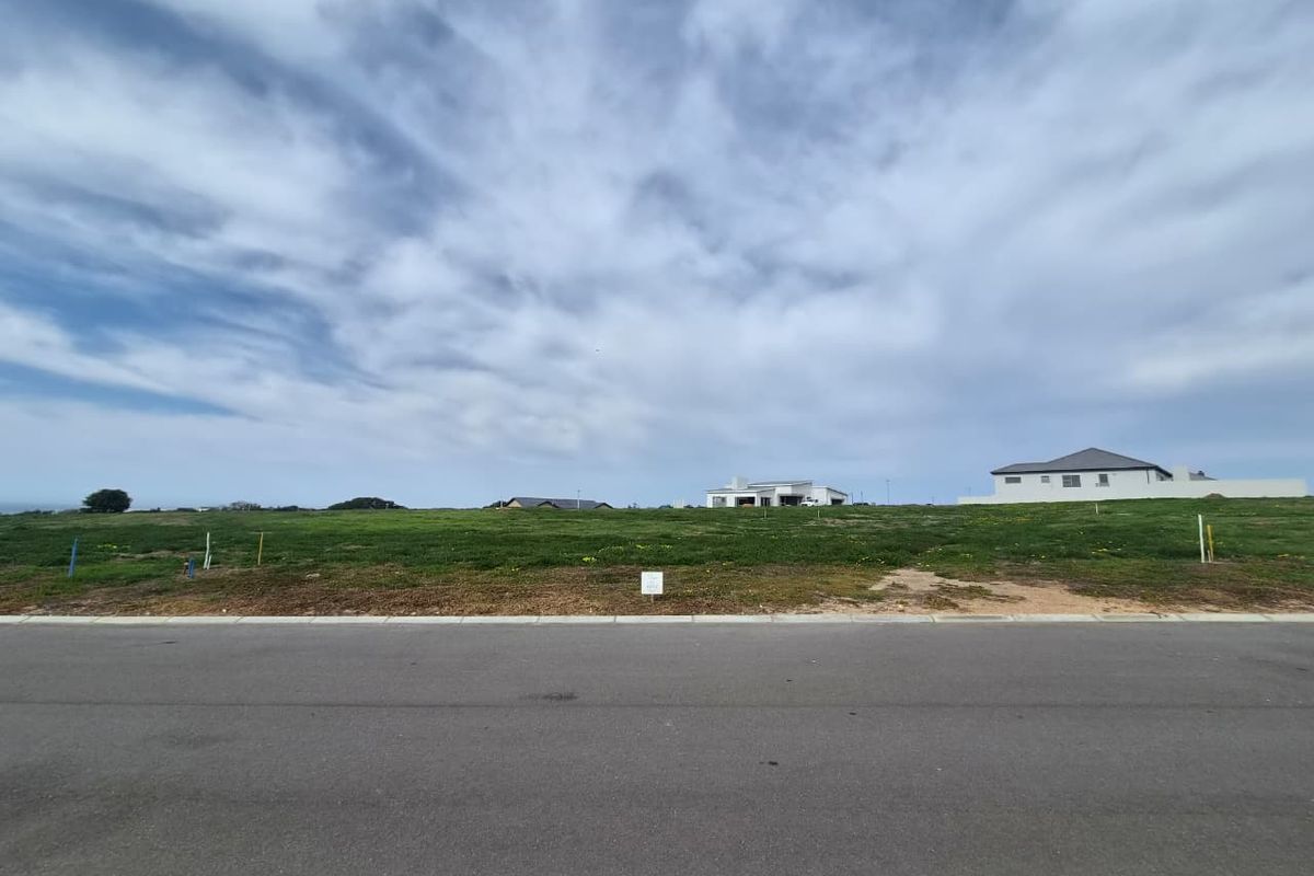 A vacant residential plot of land featuring a level grassy surface, accessible from a paved street in a developing neighborhood.