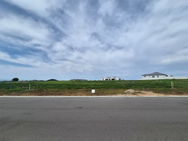 A vacant residential plot of land featuring a level grassy surface, accessible from a paved street in a developing neighborhood.