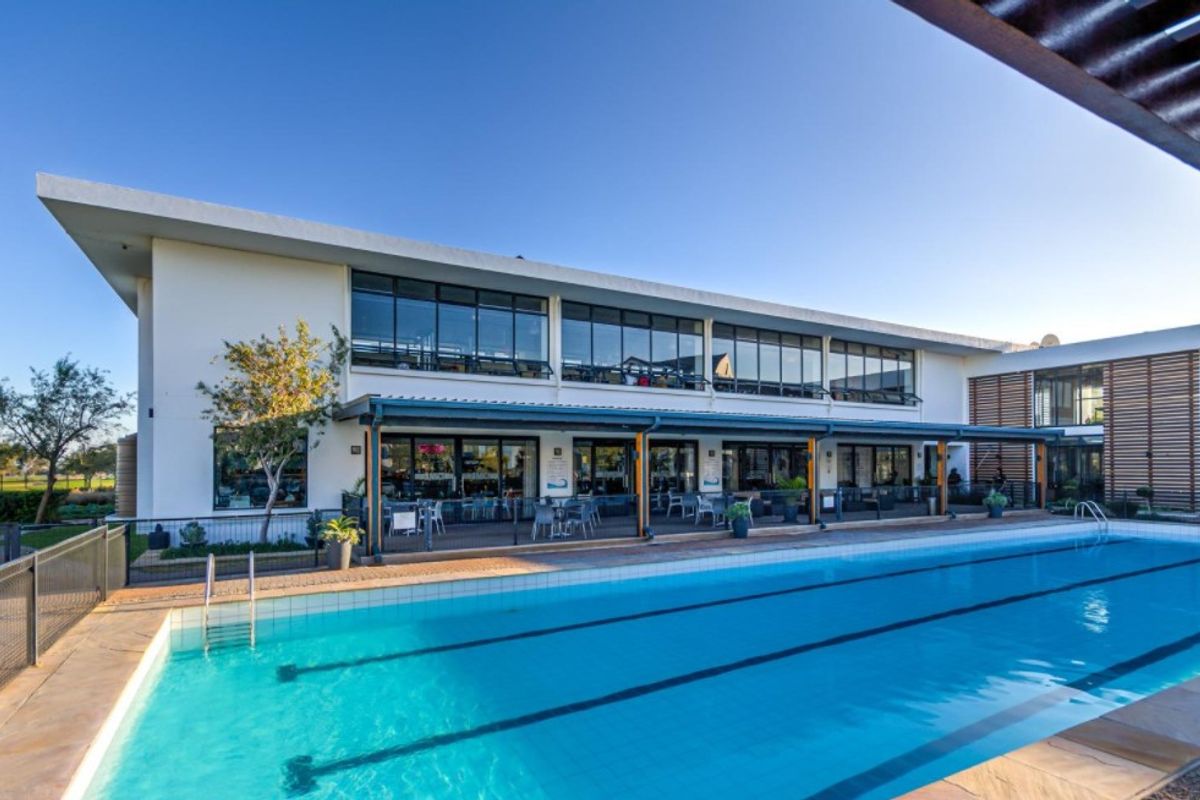 A well-maintained swimming pool area featuring a large lap pool, paved deck, and a covered patio attached to a modern multi-story building.