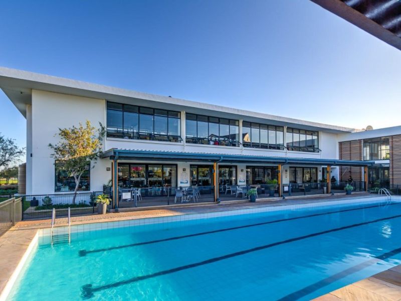 A well-maintained swimming pool area featuring a large lap pool, paved deck, and a covered patio attached to a modern multi-story building.