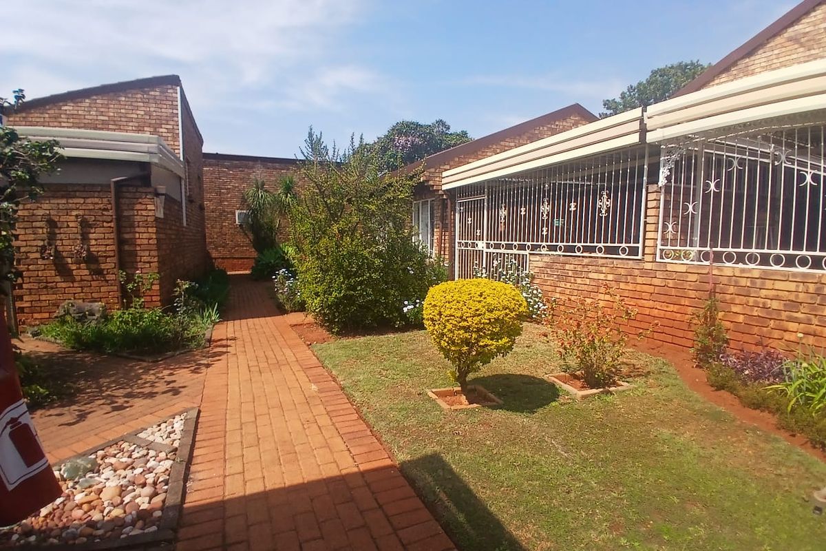 A rear exterior view of a townhouse featuring a face brick facade, a paved walkway, and a small landscaped garden area with security bars.