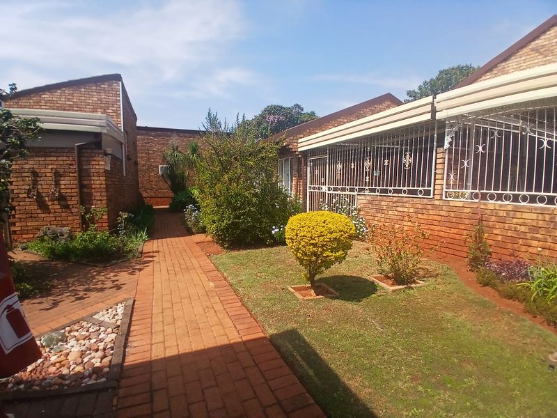 A rear exterior view of a townhouse featuring a face brick facade, a paved walkway, and a small landscaped garden area with security bars.