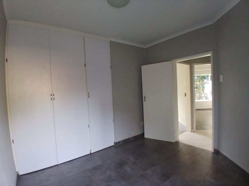 A vacant bedroom featuring built-in cupboards, neutral grey walls, and dark tiled flooring, with a doorway leading to an adjacent room.